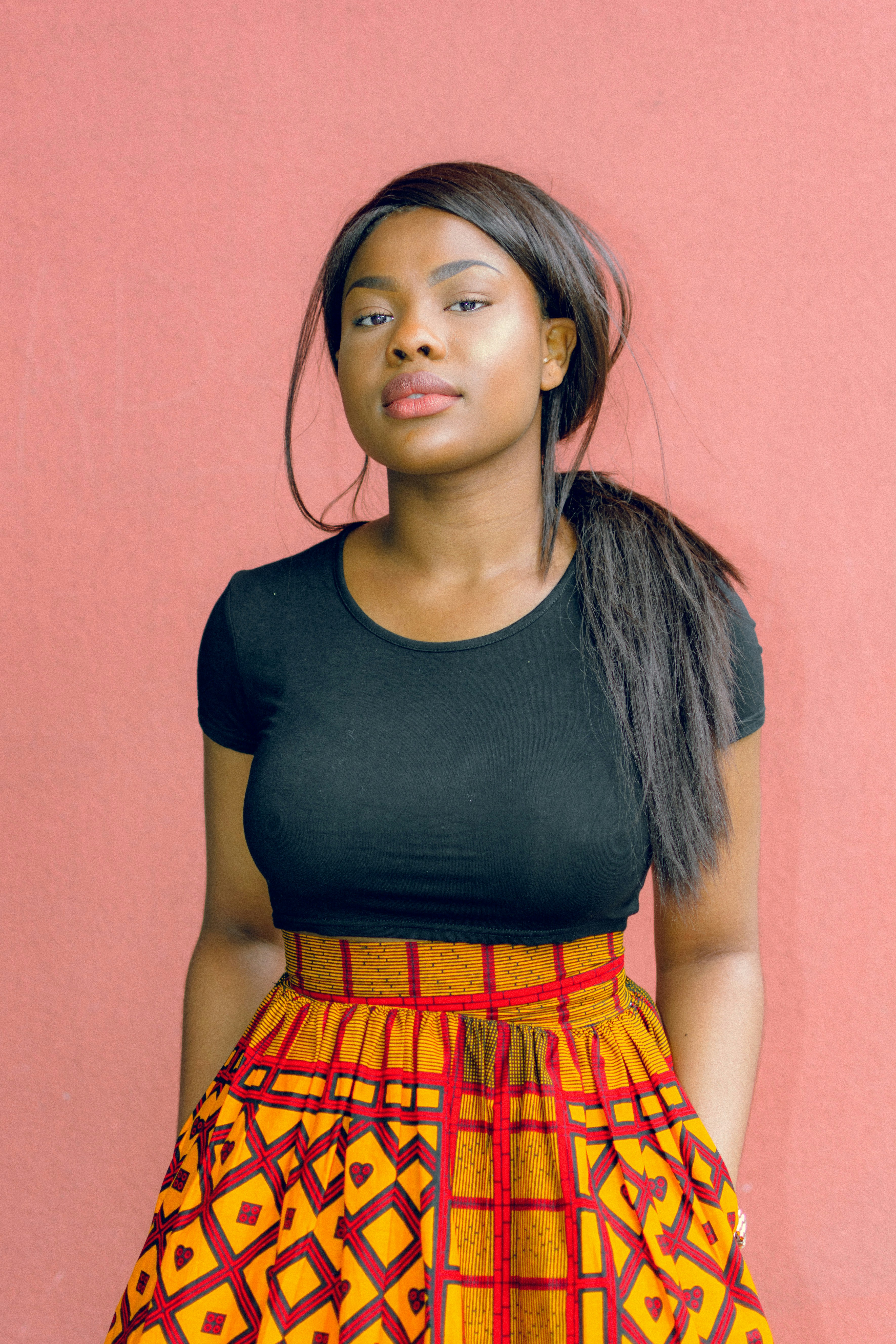 Woman standing against a pink background, wearing a black top and a vibrant orange patterned skirt, looking confident and composed.