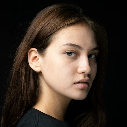 Young woman with long brown hair and neutral expression, gazing sideways against a dark background. The image conveys a calm and contemplative mood.
